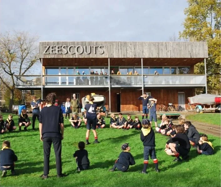 Leden zitten in een grote kring op het gras voor het zeescoutsgebouw aan de Thonetlaan